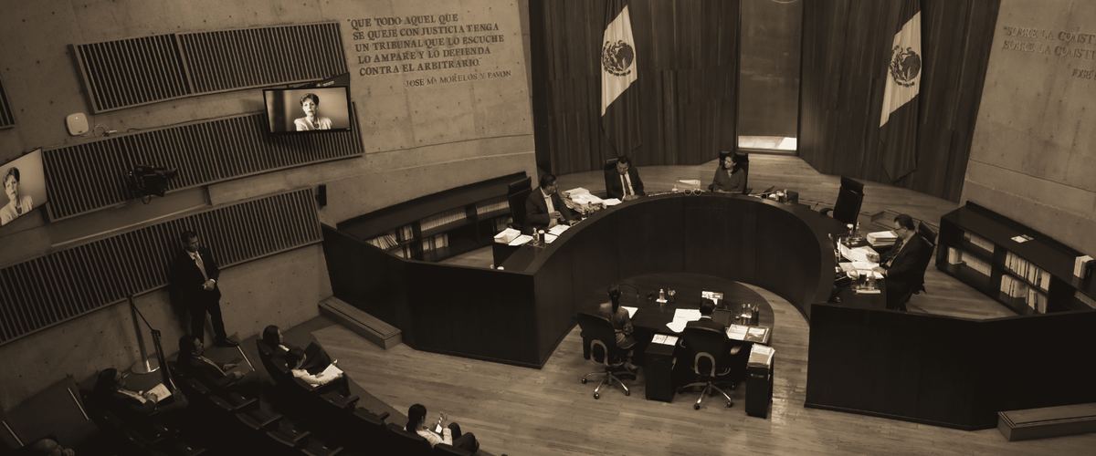 Vista de una sala de sesiones de un tribunal, con el estrado de madera y banderas de México